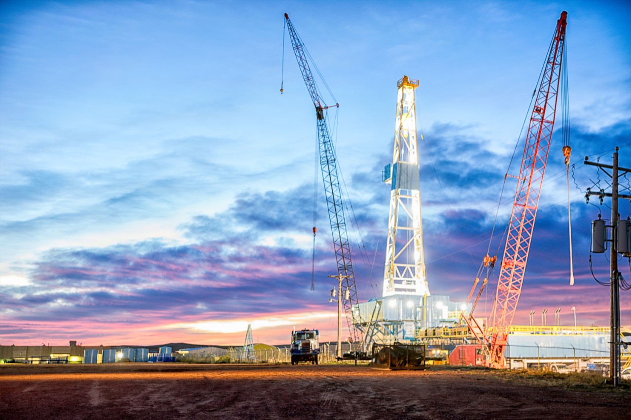 Well lit oil drilling Fracking Rig surrounded by green grass and bushes.  There are some clouds in the distance and lights on the Fracking Right side of the Fracking Rig.