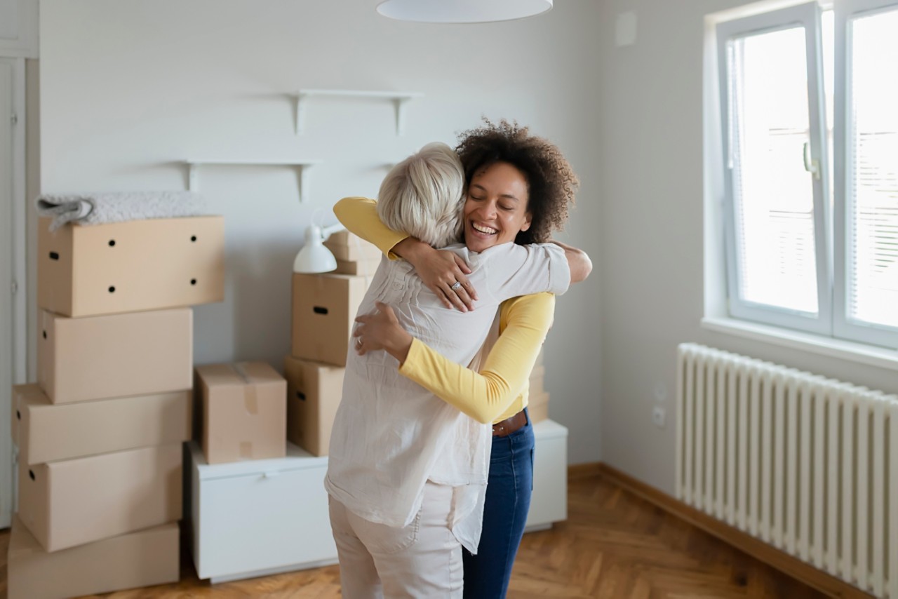 Senior Mom and Her Adopted Mixed Race Adult Daughter First Time Home Buyers Celebrate and Hugging New House Purchase on Moving Day, Laughing Standing Among Boxes in Own Flat House
