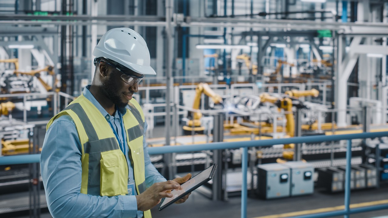 Happy African American Car Factory Engineer in High Visibility Vest Using Tablet Computer. Automotive Industrial Facility Working on Vehicle Production on Automated Technology Assembly Plant.